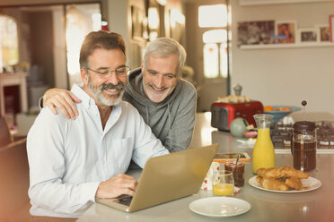 Male gay couple using laptop and eating breakfast at kitchen counter - CAIF05737