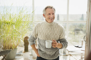 Lächelnder reifer Mann trinkt Kaffee auf der Veranda eines sonnigen Strandhauses - CAIF05750