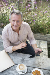 Portrait smiling senior man using digital tablet and drinking coffee at patio table - CAIF05759