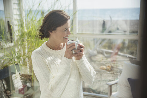 Lächelnde reife Frau trinkt Kaffee auf der Sonnenveranda eines Strandhauses - CAIF05762