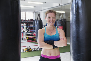 Porträt einer lächelnden, selbstbewussten weiblichen Boxerin, die in einem Fitnessstudio an Boxsäcken steht - CAIF05778