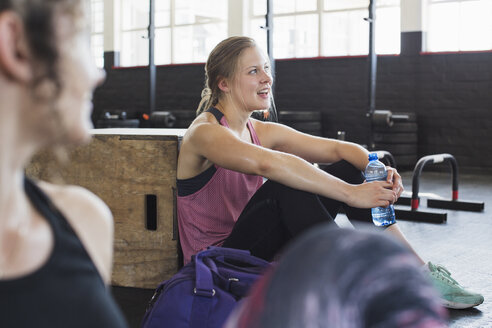 Lächelnde junge Frau ruht sich aus und trinkt Wasser nach dem Training im Fitnessstudio - CAIF05788