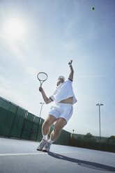 Junger männlicher Tennisspieler, der auf einem sonnigen Tennisplatz den Ball aufschlägt - CAIF05824