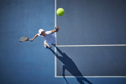 Draufsicht auf einen jungen männlichen Tennisspieler, der auf einem sonnigen blauen Tennisplatz den Ball aufschlägt - CAIF05840