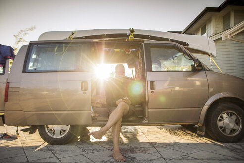 Barefoot man sitting in minivan in backlight using cell phone - GUSF00406