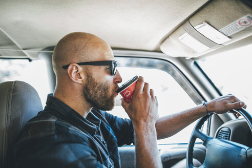 Junger Mann mit Sonnenbrille und Bart auf einer Autofahrt mit Getränk zum Mitnehmen - GUSF00408