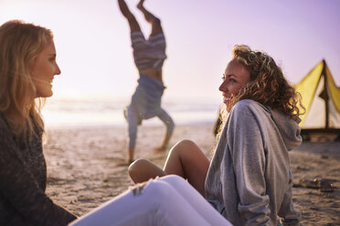 Lächelnde Frauen unterhalten sich am Strand mit einem Mann, der im Hintergrund einen Handstand macht - CAIF05874