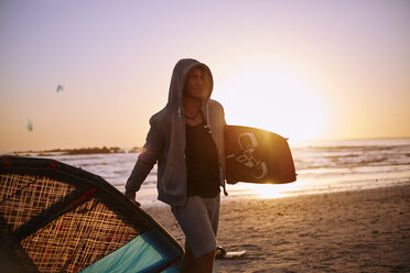 Mann in Kapuzenjacke mit Kiteboard-Ausrüstung am Strand bei Sonnenuntergang - CAIF05877