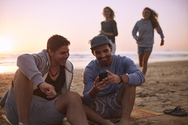 Freunde hängen mit Handy am Strand bei Sonnenuntergang herum und schreiben SMS - CAIF05881