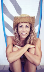 Portrait smiling female surfer in straw hat sitting in front of surfboard - CAIF05887