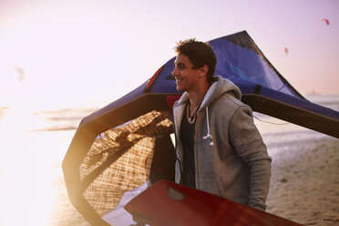 Lächelnder Mann mit Kiteboarding-Ausrüstung am Strand bei Sonnenuntergang - CAIF05898