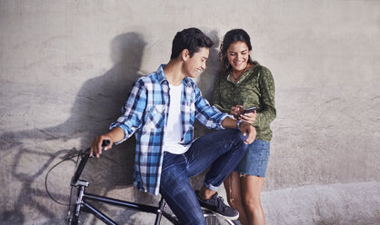 Teenage couple with BMX bicycle texting with cell phone at wall - CAIF05942