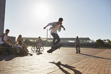 Freunde beobachten einen Teenager, der auf einem Skateboard in einem sonnigen Skatepark flippt - CAIF05945