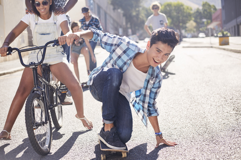 Porträt lächelnd Teenager Junge Skateboarding mit Freunden auf sonnigen städtischen Straße, lizenzfreies Stockfoto