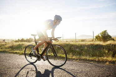 Männlicher Triathlet, der auf einer sonnigen Landstraße bei Sonnenaufgang radelt - CAIF06014