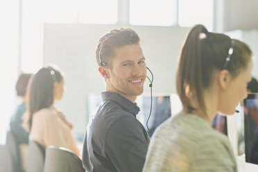 Portrait lächelnder männlicher Telefonverkäufer mit Headset am Computer in einem sonnigen Büro - CAIF06042