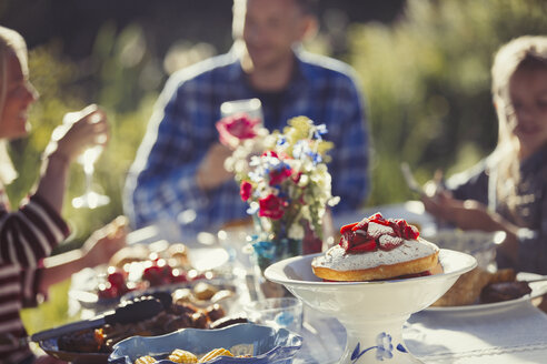 Familie genießt Erdbeerkuchen an einem sonnigen Gartenparty-Tisch im Innenhof - CAIF06061