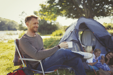 Lächelnder Vater trinkt Kaffee vor dem Zelt auf dem Campingplatz, lizenzfreies Stockfoto