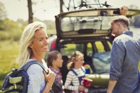 Lächelnde Frau mit Rucksack, die sich auf eine Wanderung mit ihrer Familie außerhalb des Autos vorbereitet, lizenzfreies Stockfoto