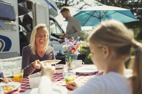 Lächelnde Mutter und Tochter genießen das Frühstück am Tisch vor dem sonnigen Wohnmobil, lizenzfreies Stockfoto