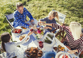 Familie stößt mit Wein- und Wassergläsern am sonnigen Mittagstisch im Innenhof an - CAIF06126