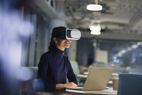 Geschäftsfrau mit Virtual-Reality-Simulator am Laptop in einem dunklen Büro, lizenzfreies Stockfoto