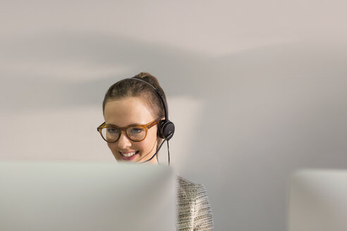 Smiling female telemarketer wearing headset talking on telephone at computer in office - CAIF06215