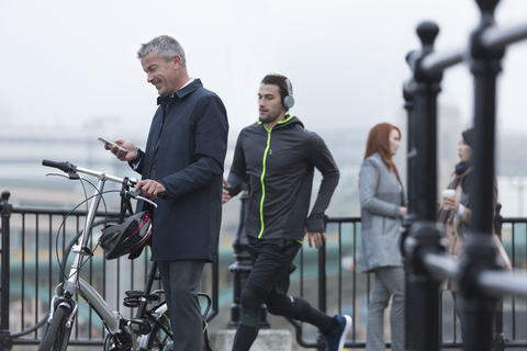 Geschäftsmann mit Fahrrad, der mit seinem Handy SMS schreibt, und männlicher Läufer auf städtischer Rampe, lizenzfreies Stockfoto