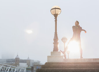 Male runner stretching arms at sunny urban lamppost - CAIF06319