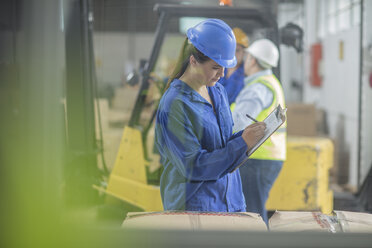 Woman wearing hard hat writing on clipboard in factory - ZEF15116