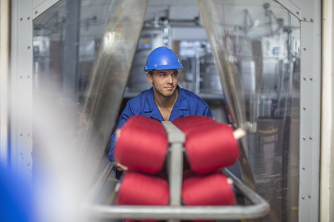 Lächelnde Frau mit Schutzhelm, die einen Wagen in einer Fabrik schiebt, lizenzfreies Stockfoto