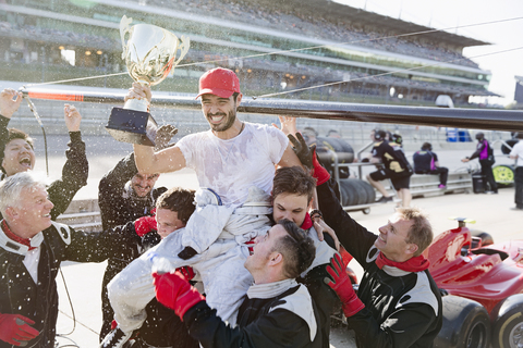 Formel-1-Rennteam, das den Fahrer mit der Trophäe auf den Schultern trägt und den Sieg auf der Sportstrecke feiert, lizenzfreies Stockfoto