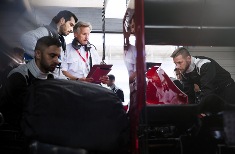 Manager und Fahrer unterhalten sich hinter der Boxencrew, die in der Reparaturwerkstatt an einem Rennwagen arbeitet, lizenzfreies Stockfoto