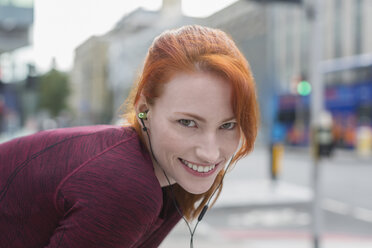 Porträt einer lächelnden Läuferin mit Kopfhörern, die sich auf einem Bürgersteig ausruht - CAIF06555