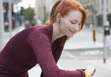 Lächelnde Läuferin mit roten Haaren und Kopfhörern, die ihre Smartwatch überprüft - CAIF06558