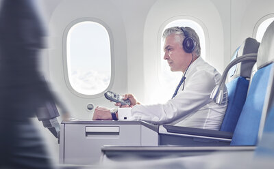 Geschäftsmann mit Kopfhörern und Fernbedienung sieht sich im Flugzeug einen Film an - CAIF06603
