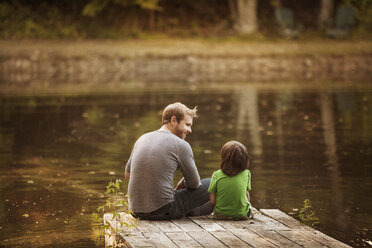 Rear view of father and son sitting on jetty by lake - CAVF01211