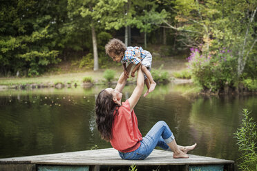 Glückliche Frau hebt ihren Sohn hoch, während sie auf einem Steg am See sitzt - CAVF01246