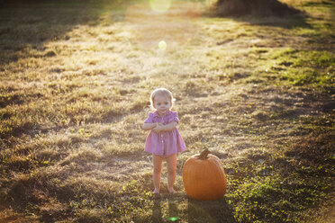 Cute baby girl standing by pumpkin on grassy field - CAVF01296