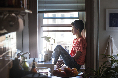Frau schaut weg, während sie zu Hause auf der Fensterbank sitzt, lizenzfreies Stockfoto