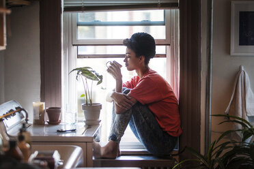 Side view of woman drinking drink while sitting on window sill at home - CAVF01308