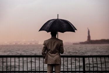 Rückansicht eines Mannes mit Regenschirm, der an einer Reling am Meer steht - CAVF01336