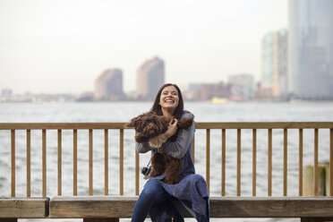 Playful woman with dog sitting on bench against Hudson River - CAVF01346