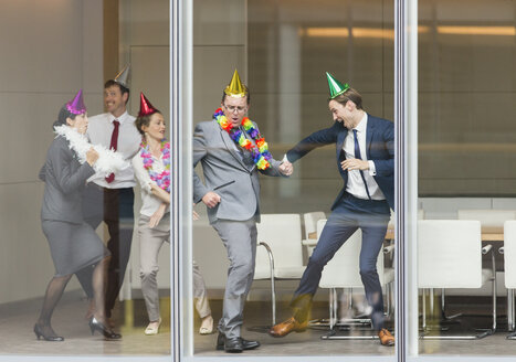 Verspielte Geschäftsleute mit Partyhüten tanzen am Fenster des Konferenzraums - CAIF06616