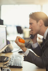 Angry businessman squeezing stress ball and talking on telephone at computer in office - CAIF06675