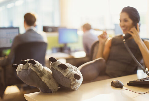 Geschäftsfrau in Wolfspfoten-Pantoffeln mit hochgelegten Füßen auf dem Schreibtisch beim Telefonieren - CAIF06676
