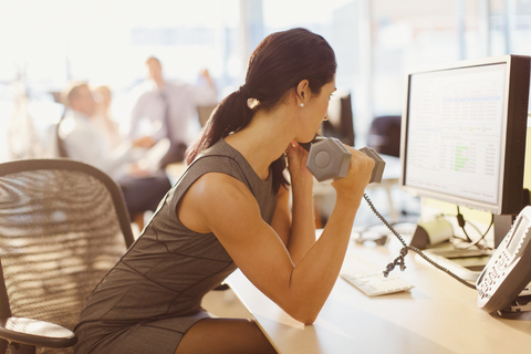 Businesswoman doing biceps curls with dumbbell and talking on telephone at computer in office stock photo