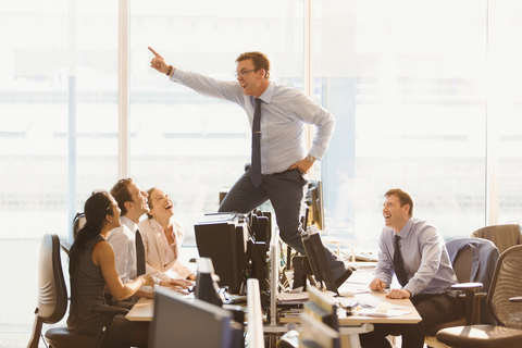 Laughing colleagues watching exuberant businessman dancing on top of desks in office stock photo