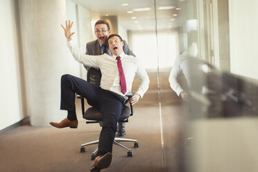 Playful businessman pushing colleague down corridor in office chair - CAIF06694