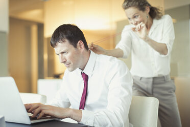Mischievous businesswoman aiming rubber band at unsuspecting businessman working at laptop - CAIF06700